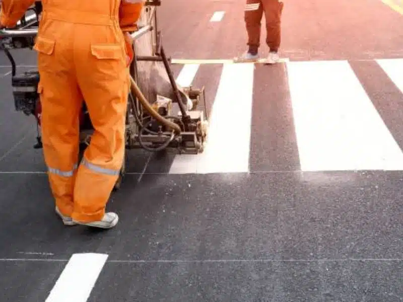 Road construction crew applying fresh white crosswalk striping on asphalt with pavement marking equipment.