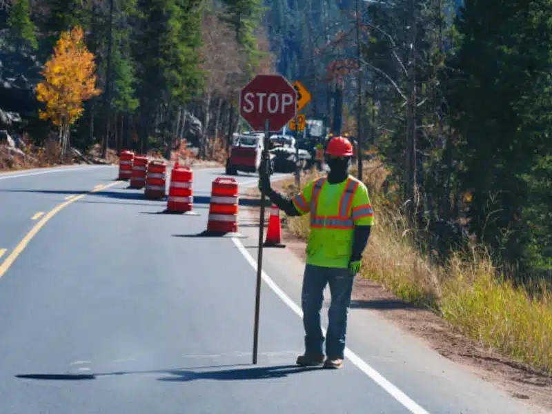 A construction worker in a neon safety vest and helmet holds a stop sign on a two-lane road lined with orange traffic barrels, surrounded by trees and a forested landscape.