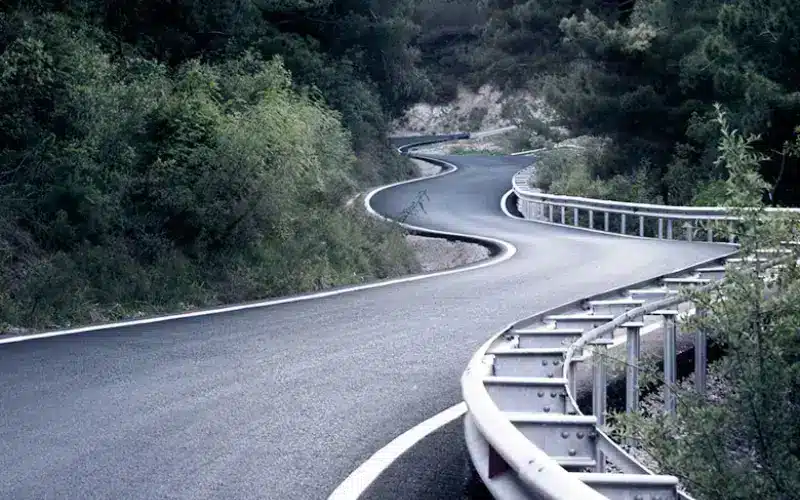 Curving mountain road with fresh asphalt and metal guardrails winding through dense green trees.